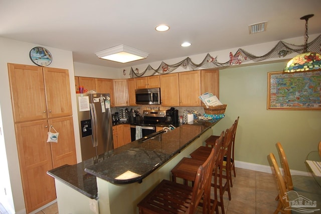 kitchen with light tile patterned floors, a breakfast bar, appliances with stainless steel finishes, dark stone countertops, and kitchen peninsula