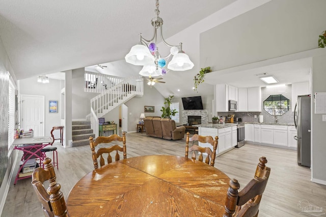 dining space featuring high vaulted ceiling, a ceiling fan, a stone fireplace, light wood finished floors, and stairs