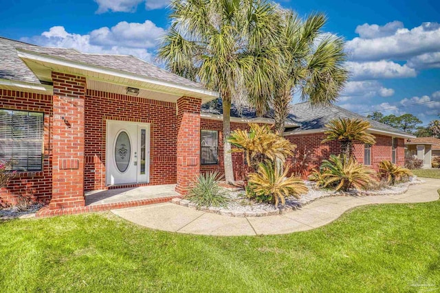 doorway to property with a yard, brick siding, and roof with shingles