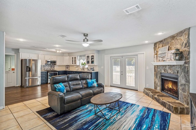 living room featuring ceiling fan, a textured ceiling, light tile patterned flooring, a stone fireplace, and french doors