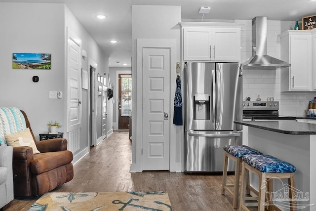 kitchen with white cabinetry, wall chimney range hood, stainless steel appliances, dark hardwood / wood-style flooring, and a breakfast bar area