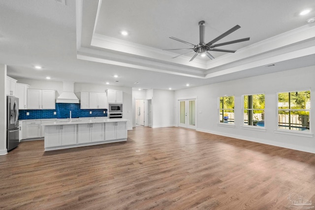 kitchen featuring appliances with stainless steel finishes, custom range hood, wood-type flooring, a center island with sink, and white cabinetry