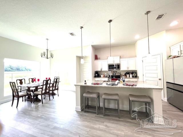 kitchen with light wood-type flooring, decorative light fixtures, appliances with stainless steel finishes, a center island with sink, and white cabinets