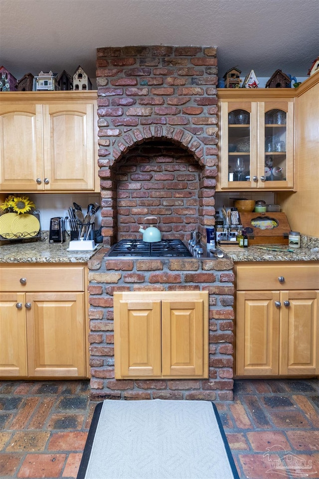 kitchen featuring stainless steel gas stovetop, glass insert cabinets, brick floor, and light brown cabinets