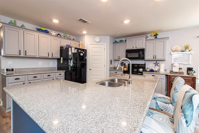 kitchen with light stone counters, sink, a center island with sink, black appliances, and a breakfast bar