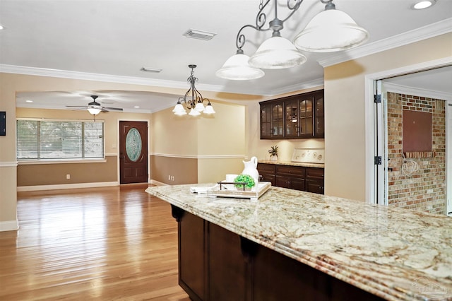 kitchen with pendant lighting, dark brown cabinets, light stone counters, ornamental molding, and light wood-type flooring