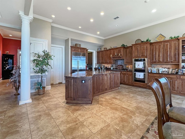 kitchen with a kitchen island with sink, appliances with stainless steel finishes, ornamental molding, tasteful backsplash, and ornate columns