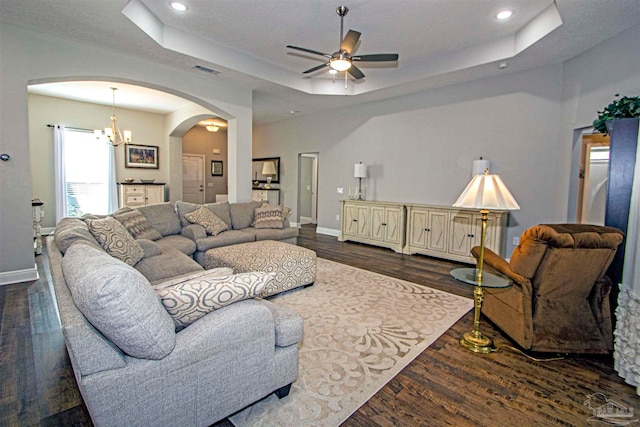 living room with ceiling fan with notable chandelier, a raised ceiling, and dark wood-type flooring