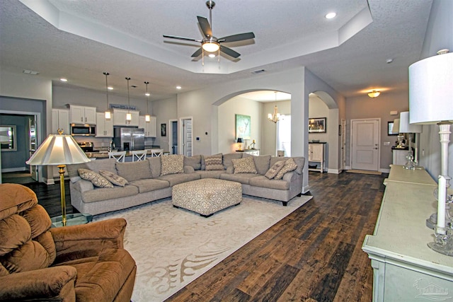 living room with ceiling fan with notable chandelier, a textured ceiling, a tray ceiling, and dark wood-type flooring