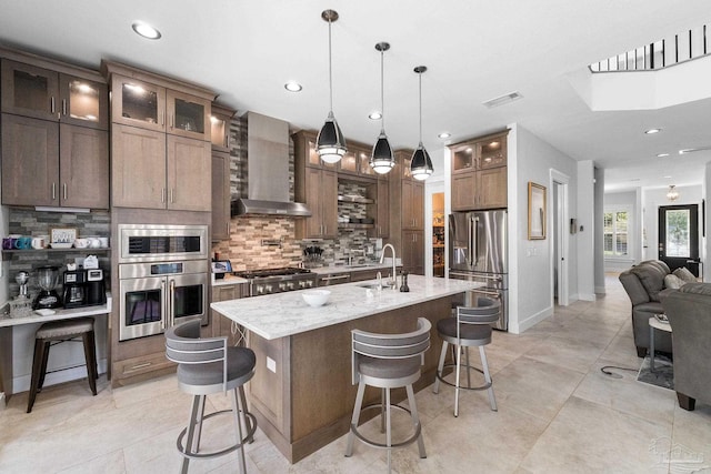 kitchen featuring appliances with stainless steel finishes, hanging light fixtures, an island with sink, light stone counters, and wall chimney range hood