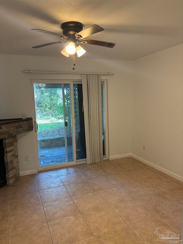 empty room featuring a textured ceiling, light tile patterned floors, a stone fireplace, and ceiling fan