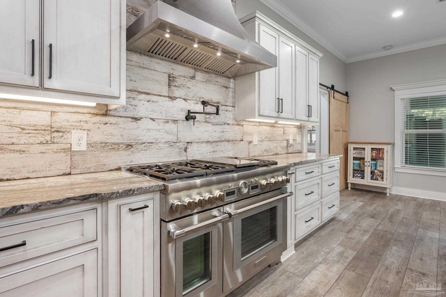 kitchen featuring range with two ovens, light hardwood / wood-style floors, wall chimney range hood, white cabinetry, and a barn door