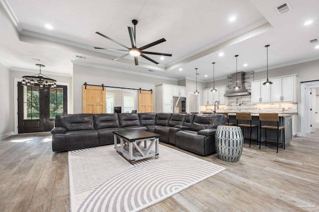 living room featuring a barn door, ceiling fan with notable chandelier, light hardwood / wood-style flooring, a raised ceiling, and ornamental molding