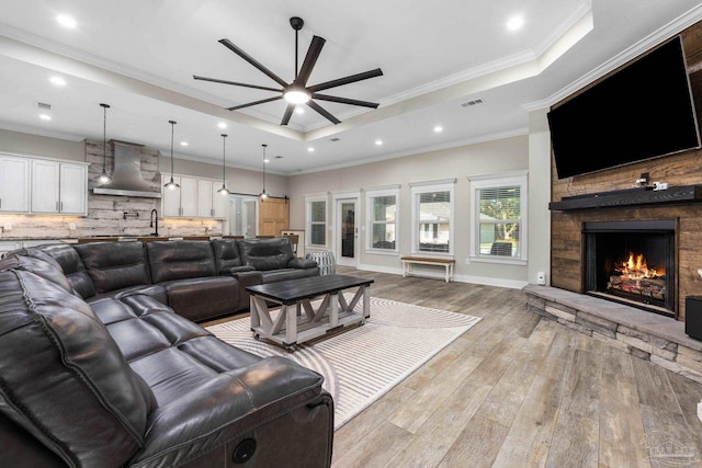 living room featuring ceiling fan, a raised ceiling, ornamental molding, light hardwood / wood-style flooring, and a fireplace