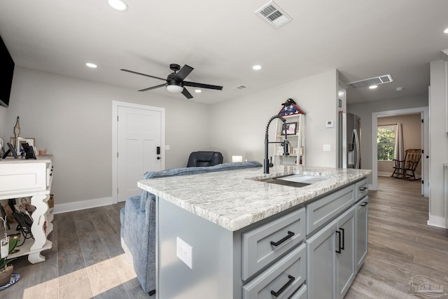 kitchen with a kitchen island with sink, ceiling fan, light stone counters, sink, and hardwood / wood-style flooring