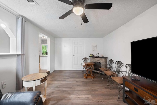 living area featuring ceiling fan, a stone fireplace, dark hardwood / wood-style floors, and a textured ceiling