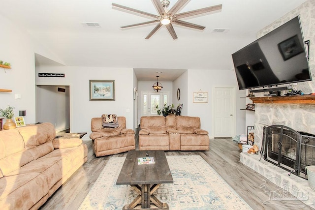 living room featuring light wood-type flooring, ceiling fan with notable chandelier, and a stone fireplace