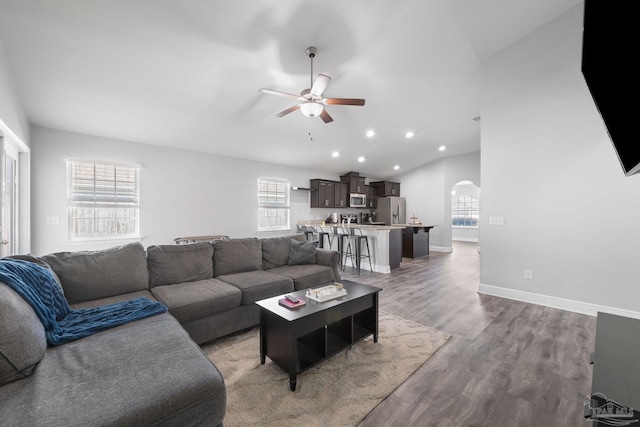 living area featuring a ceiling fan, wood finished floors, baseboards, lofted ceiling, and arched walkways