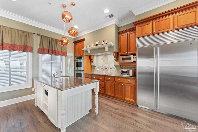 kitchen with an island with sink, brown cabinets, dark stone countertops, stainless steel appliances, and pendant lighting