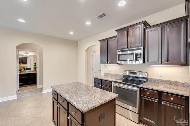 kitchen featuring light tile patterned floors, light stone counters, dark brown cabinets, a kitchen island, and appliances with stainless steel finishes