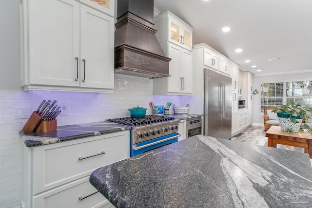 kitchen with tasteful backsplash, dark stone counters, high end appliances, white cabinets, and premium range hood