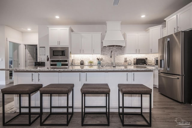 kitchen with custom range hood, appliances with stainless steel finishes, a center island with sink, and white cabinets