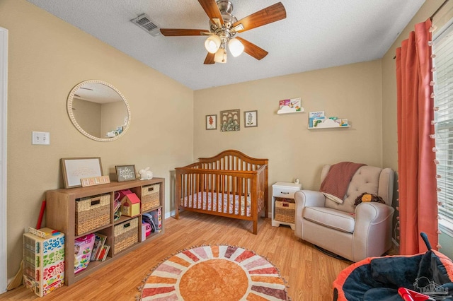 bedroom with a nursery area, a textured ceiling, visible vents, a ceiling fan, and wood finished floors