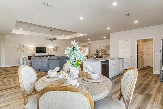 dining room featuring sink, ceiling fan, a textured ceiling, a tray ceiling, and light wood-type flooring