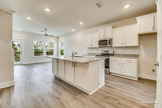 kitchen with light stone countertops, stainless steel appliances, a kitchen island with sink, white cabinets, and light hardwood / wood-style floors