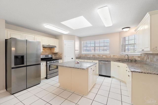 kitchen with light tile patterned floors, under cabinet range hood, a sink, appliances with stainless steel finishes, and decorative backsplash