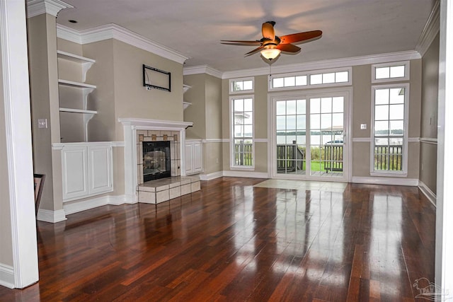 unfurnished living room with ceiling fan, built in shelves, a tile fireplace, crown molding, and dark hardwood / wood-style flooring