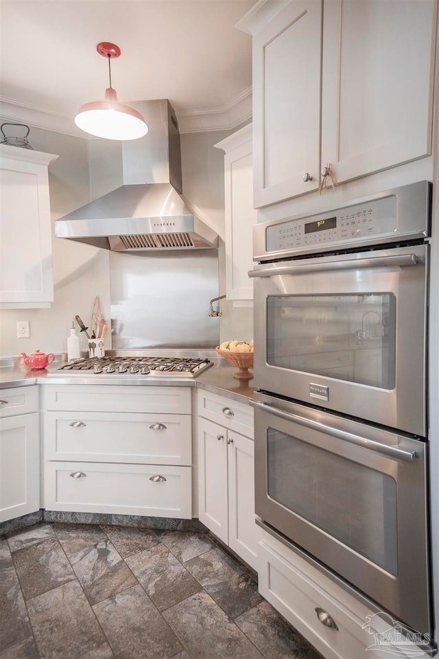 kitchen featuring wall chimney range hood, appliances with stainless steel finishes, hanging light fixtures, white cabinetry, and ornamental molding