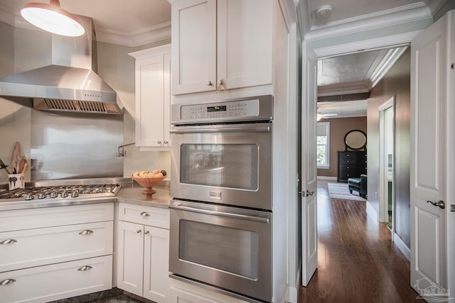 kitchen featuring wall chimney range hood, dark wood-type flooring, stainless steel appliances, ornamental molding, and white cabinets