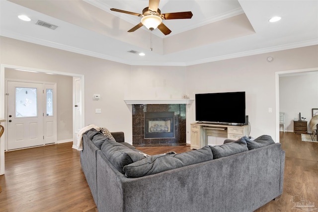 living room featuring ceiling fan, a tiled fireplace, a tray ceiling, ornamental molding, and dark hardwood / wood-style flooring