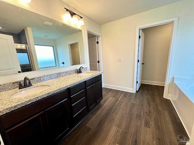 full bath with wood finished floors, double vanity, a garden tub, a textured ceiling, and baseboards