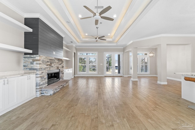 unfurnished living room with ornamental molding, ceiling fan with notable chandelier, a tray ceiling, light hardwood / wood-style floors, and a stone fireplace