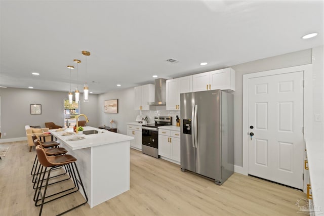 kitchen with a kitchen island with sink, wall chimney range hood, white cabinetry, appliances with stainless steel finishes, and light hardwood / wood-style floors