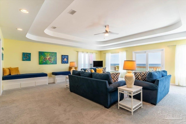 carpeted living room with a raised ceiling, a healthy amount of sunlight, visible vents, and ceiling fan