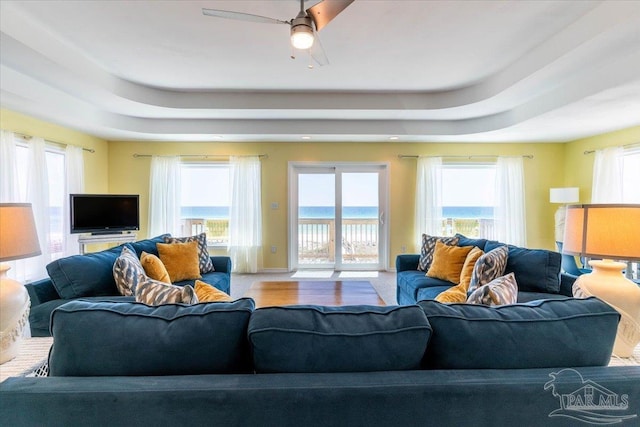 living area featuring plenty of natural light, ceiling fan, baseboards, and a tray ceiling