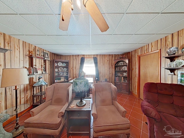 tiled living room featuring built in shelves, a paneled ceiling, ceiling fan, and wood walls