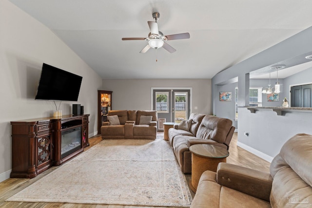 living room featuring baseboards, light wood-type flooring, vaulted ceiling, ceiling fan with notable chandelier, and a glass covered fireplace