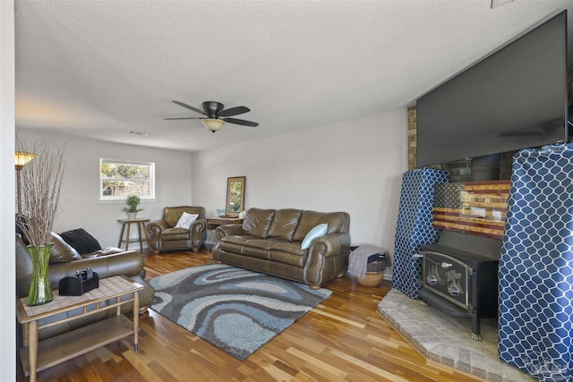 living area with ceiling fan, a textured ceiling, wood finished floors, visible vents, and a wood stove