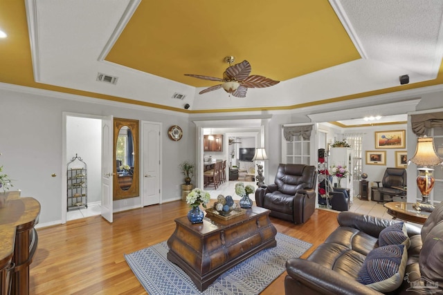 living room featuring ceiling fan, ornamental molding, a raised ceiling, and light wood-type flooring