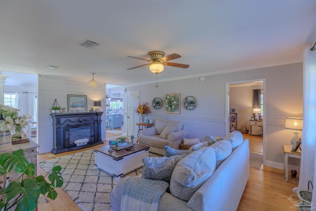 living room with crown molding, light wood-type flooring, visible vents, a ceiling fan, and a glass covered fireplace