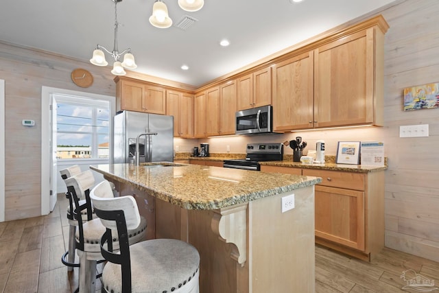 kitchen with wood walls, a kitchen island with sink, light hardwood / wood-style flooring, decorative light fixtures, and stainless steel appliances