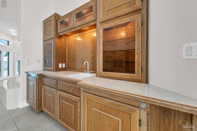 kitchen featuring light tile patterned floors, tile counters, glass insert cabinets, brown cabinets, and a sink