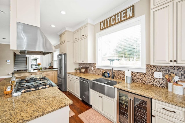 kitchen with beverage cooler, island range hood, appliances with stainless steel finishes, light stone countertops, and sink