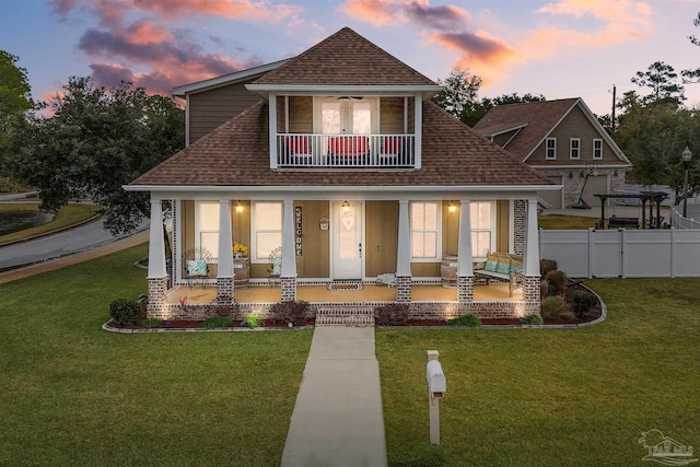 back house at dusk featuring a balcony, ceiling fan, a yard, and covered porch