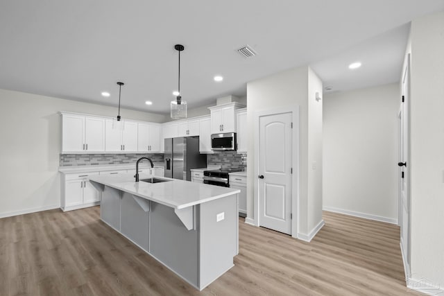 kitchen featuring light wood-type flooring, an island with sink, stainless steel appliances, decorative light fixtures, and white cabinets