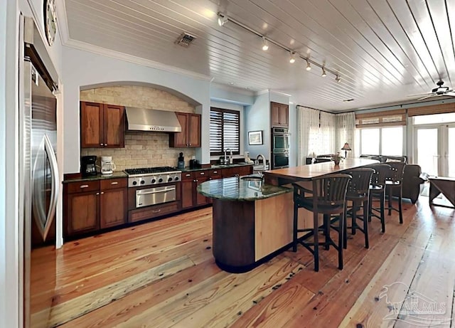 kitchen with track lighting, sink, light hardwood / wood-style floors, an island with sink, and wall chimney exhaust hood